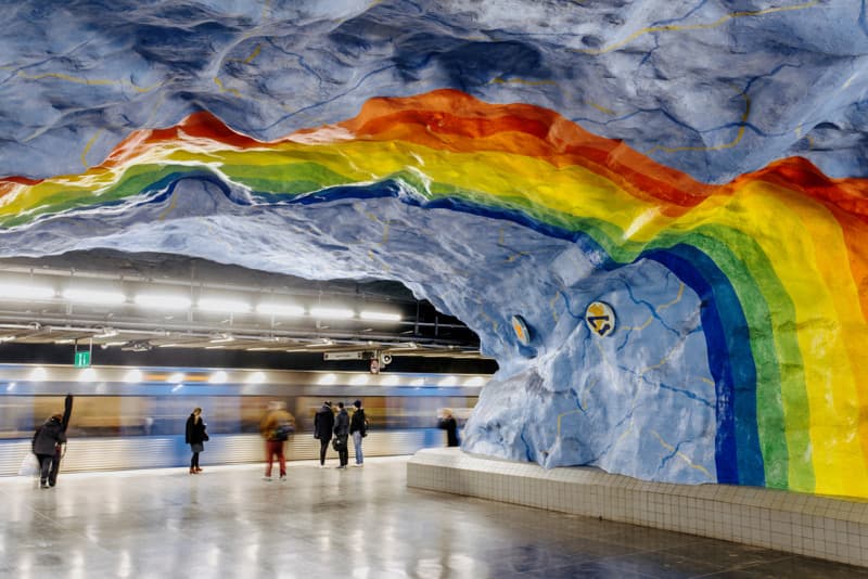 Vibrant rainbow mural painted across the blue rock wall of a metro station, with passengers waiting on the platform.