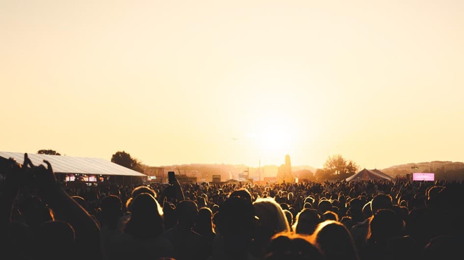 A large festival crowd stands outdoors facing the setting sun, with tents and trees silhouetted against a warm golden sky.