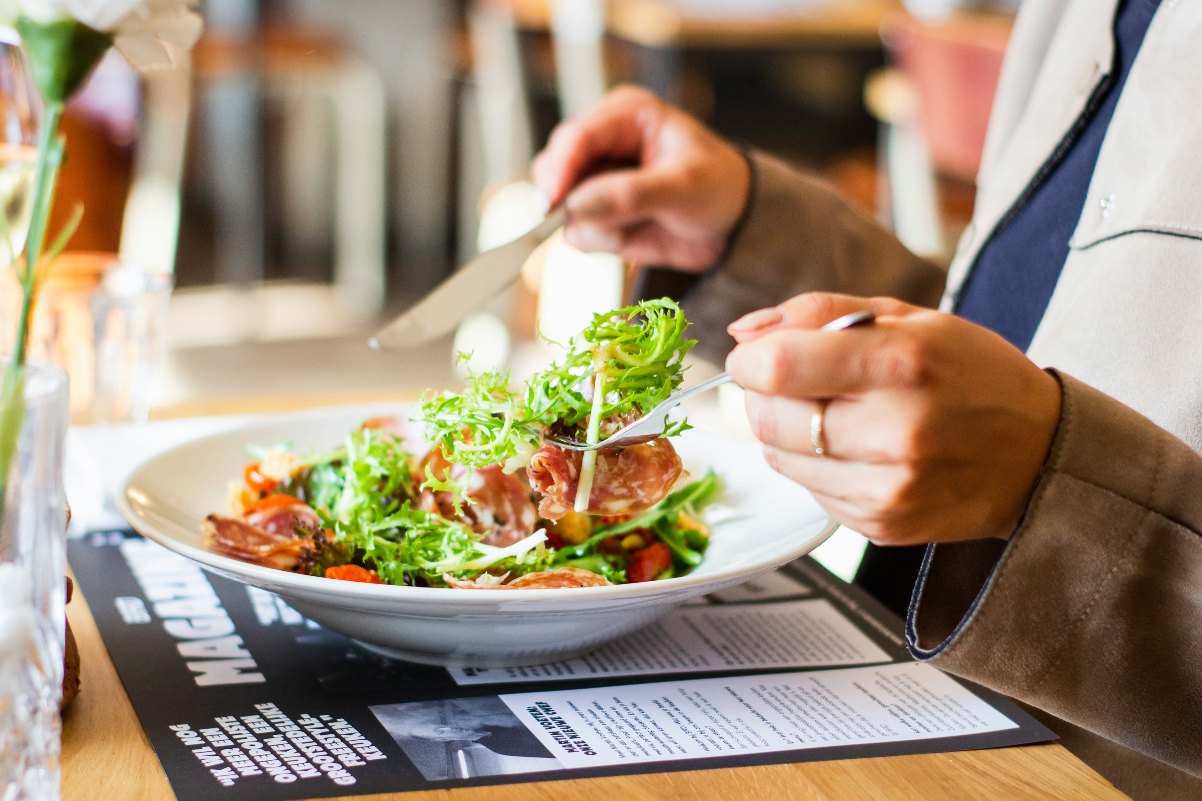 A person enjoys a fresh salad with greens and sliced meat from a white bowl at a Stockholm restaurant, with warm wooden tones in the background.