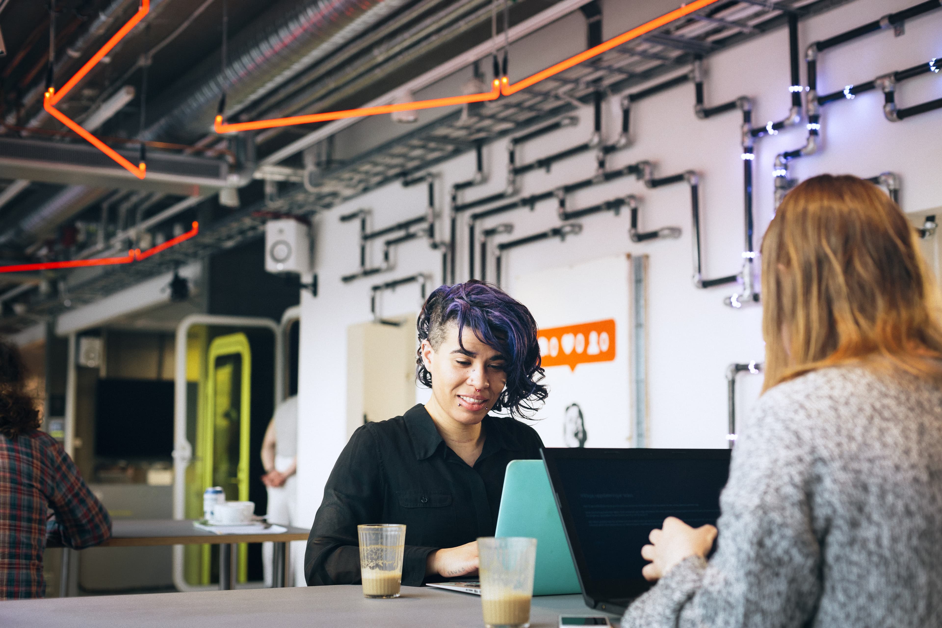 Two people sitting at a table with laptops and coffee in a modern space with neon lights and industrial decor.
