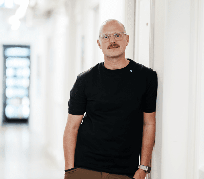 Mange Villson looking amicably at the spectator and leaning against a white wall, wearing a black t-shirt. He is a middle-aged man with a shaved head, a moustasche and transparent glasses.