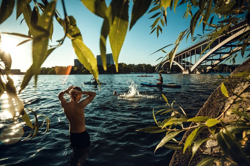 Summer swim by the bridge People swimming and paddleboarding in the evening sun near a bridge, seen through overhanging green leaves by the water.