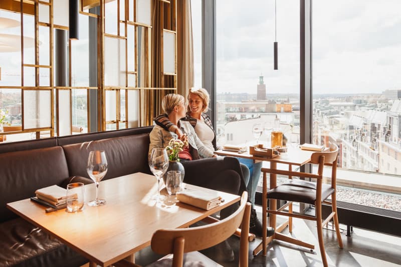 Two people sit close together on a sofa by a restaurant window, smiling at each other over set tables, with a panoramic city view and a distant city hall tower outside.